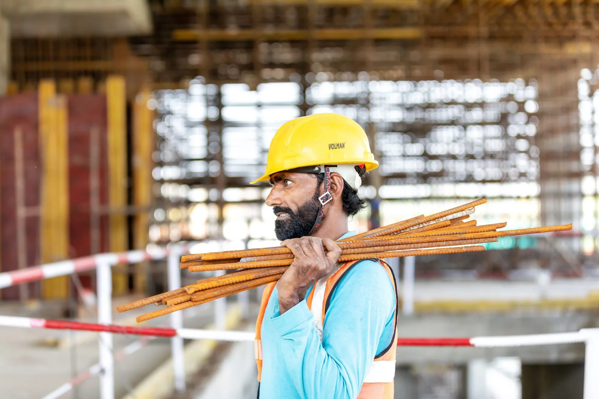 A construction worker in a hard hat and hi-vis vest carrying a bundle of reinforcing bar on one shoulder