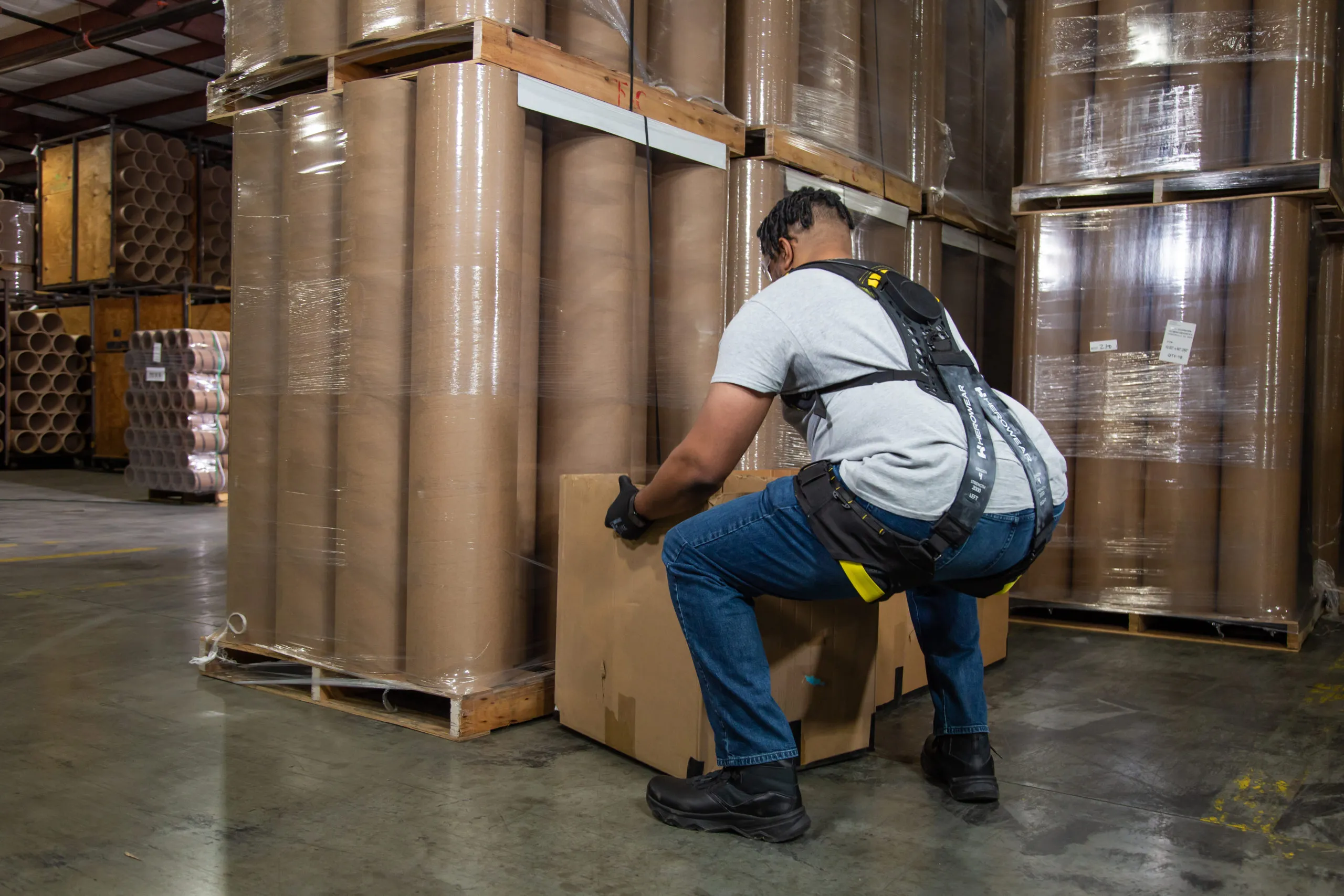 A warehouse worker wearing the Apex 2 crouching to lift a carton from a pallet in front of stacked paper rolls